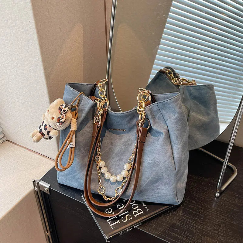 On-model cream canvas shoulder tote with beaded handle and teddy bear charm, carried at side indoors.